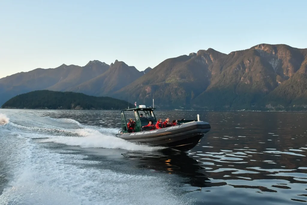 A black speedboat with several people in red jackets cruises quickly across calm water during a Whale Watching Vancouver tour, leaving a white wake behind, with forested mountains and a clear sky in the background.